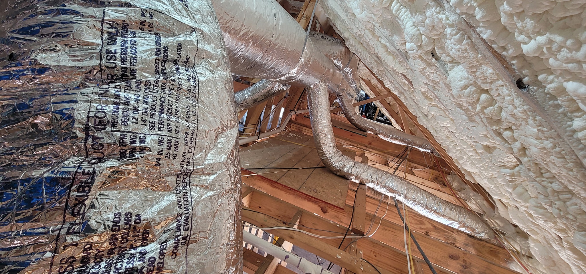 A close view shows reflective flex duct beside spray foam insulation in an attic — diagnosing HVAC short-cycling in a foam-insulated house