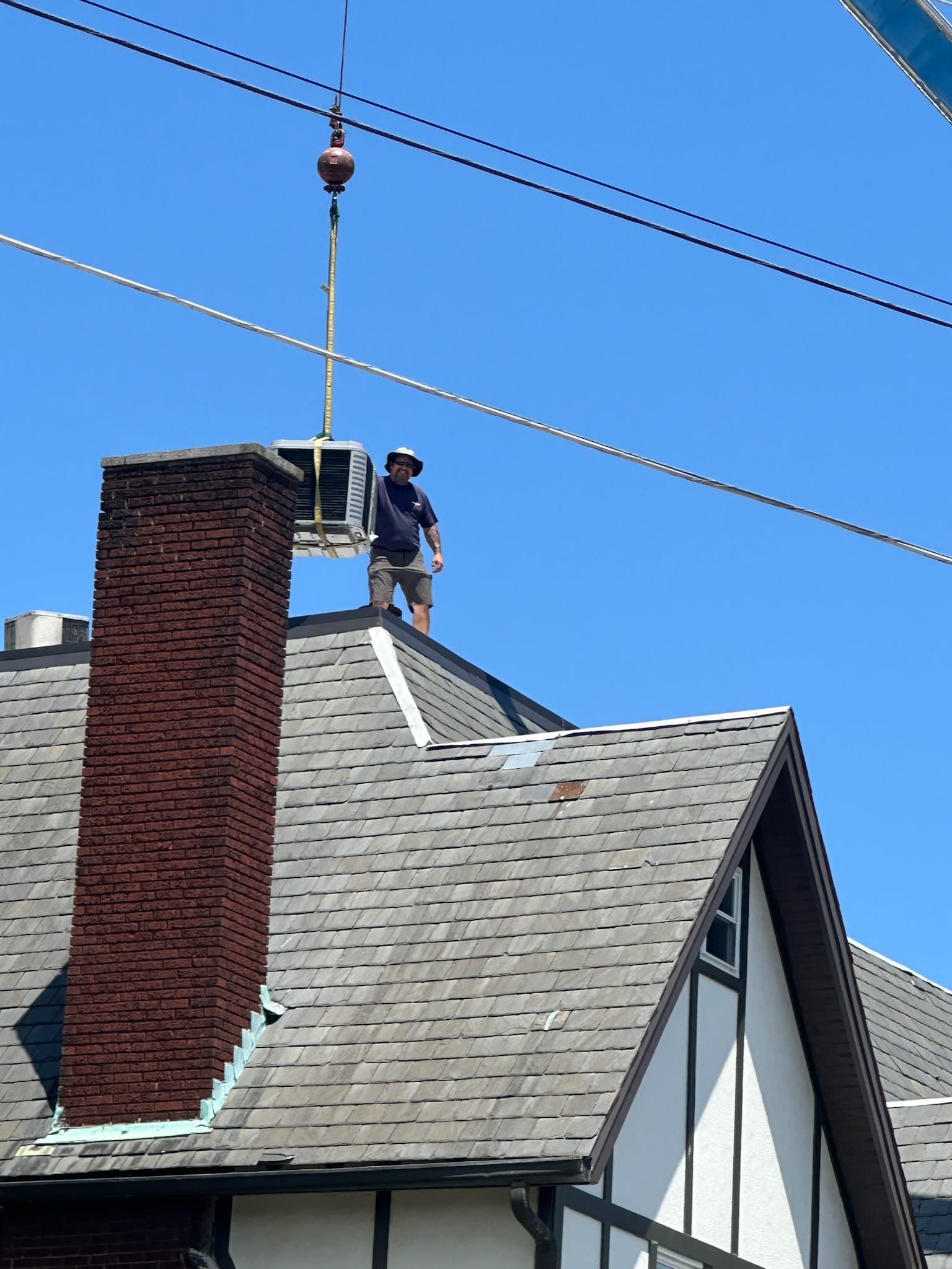 Kilgore Heating and Air crew guiding a rooftop HVAC unit into position beside a residential brick chimney